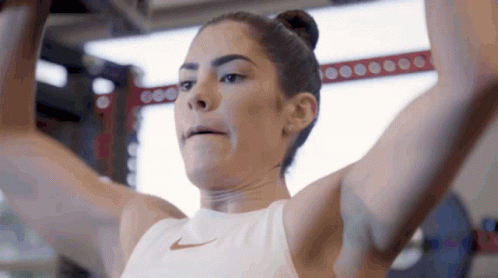 A Woman In A White Nike Tank Top Is Lifting A Barbell In A Gym GIF