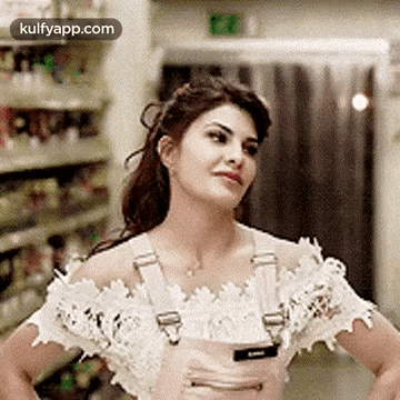 A Woman In A White Off The Shoulder Top And Pink Overalls Is Standing With Her Hands On Her Hips In A Store . GIF