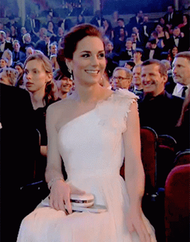 A Woman In A White One Shoulder Dress Is Smiling In Front Of A Crowd GIF