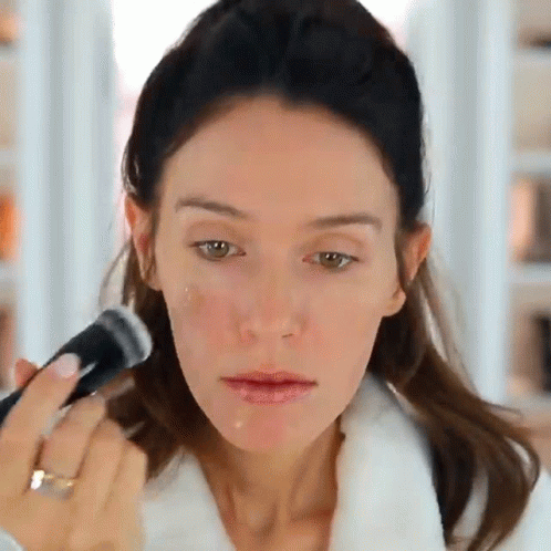 A Woman In A White Robe Is Applying Makeup To Her Face With A Brush GIF