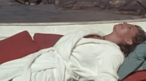 A Woman In A White Robe Is Laying On A Red Cushion On A Beach . GIF