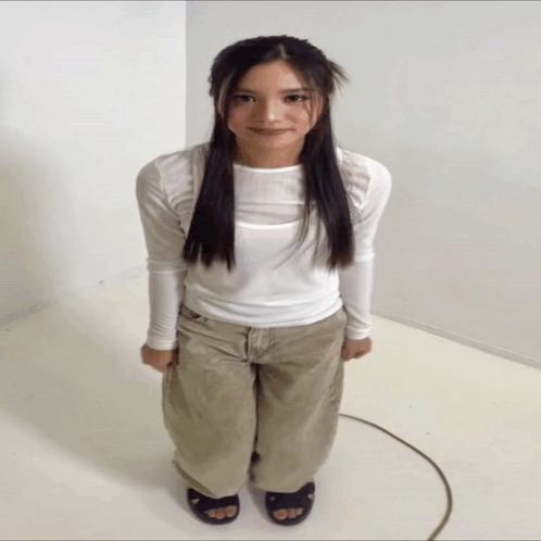 A Woman In A White Shirt And Khaki Pants Is Standing In A White Room GIF