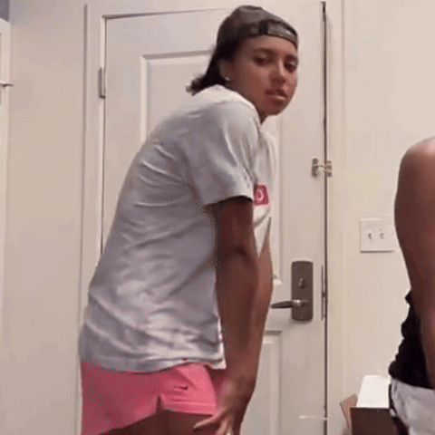 A Woman In A White Shirt And Pink Shorts Is Standing In Front Of A White Door . GIF