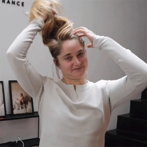 A Woman In A White Shirt Holds Her Hair In A Ponytail In Front Of A Wall That Says Rance GIF