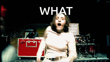 A Woman In A White Shirt Is Dancing In Front Of A Drum Set With The Word What Above Her GIF