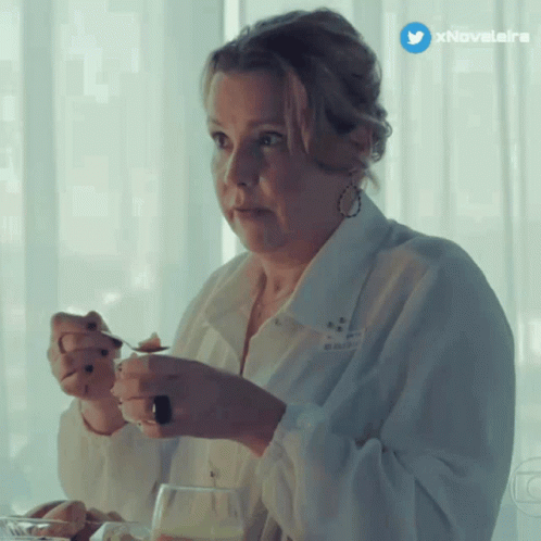 A Woman In A White Shirt Is Sitting At A Table With A Spoon In Her Hand And A Twitter Icon Above Her GIF