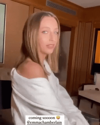 A Woman In A White Shirt Is Sitting In A Room With A Caption That Says Coming Sooooon @Emmachamberlain GIF