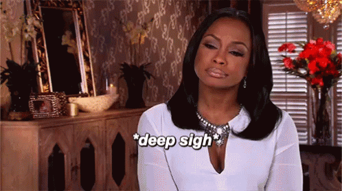 A Woman In A White Shirt Is Sitting In Front Of A Dresser And Flowers And Says Deep Sigh GIF