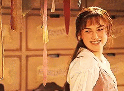 A Woman In A White Shirt Is Smiling In Front Of A Wall With Ribbons Hanging From It . GIF
