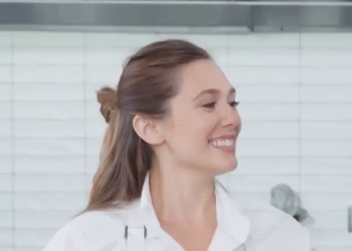 A Woman In A White Shirt Is Smiling In Front Of A White Tiled Wall . GIF