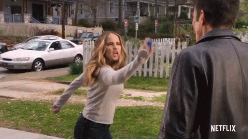 A Woman In A White Shirt Is Throwing A Bottle At A Man In A Leather Jacket . GIF