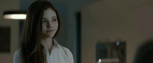 A Woman In A White Shirt Looks At The Camera In A Dark Room GIF