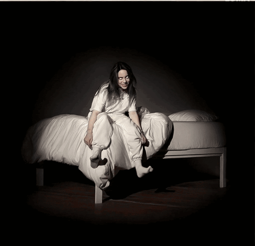 A Woman In A White Shirt Sits On A Bed GIF