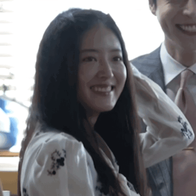 A Woman In A White Shirt With Flowers On It Smiles Next To A Man In A Suit And Tie GIF