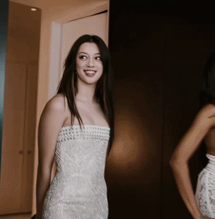A Woman In A White Strapless Dress Is Smiling In Front Of A Mirror GIF