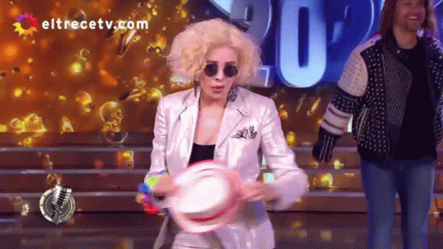 A Woman In A White Suit Is Holding A Tambourine In Front Of A Screen That Says Eltrecetv.com GIF
