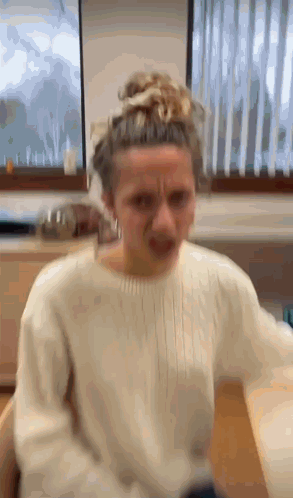 A Woman In A White Sweater Is Making A Funny Face In Front Of A Window . GIF
