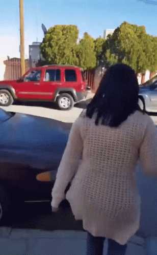 A Woman In A White Sweater Is Walking Down A Street In Front Of A Red Suv GIF