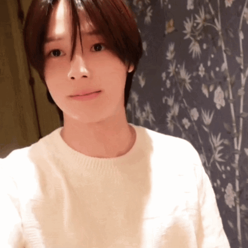 A Woman In A White Sweater Stands In Front Of A Wall With Flowers On It GIF