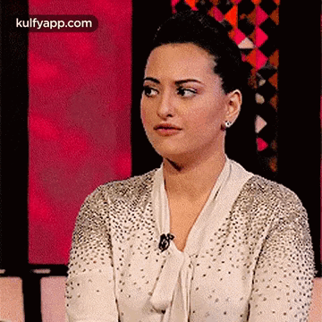 A Woman In A White Sweater With Rhinestones Is Sitting In Front Of A Pink Background . GIF