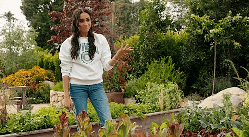 A Woman In A White Sweatshirt Is Standing In A Garden Surrounded By Plants And Flowers . GIF