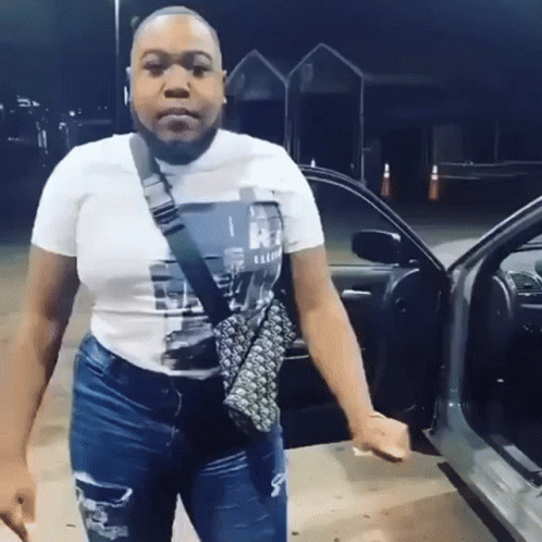A Woman In A White T-Shirt And Jeans Is Standing In Front Of A Car . GIF
