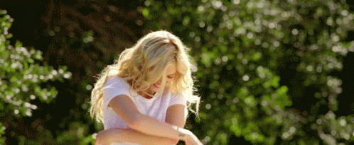 A Woman In A White T-Shirt Is Standing In Front Of Trees GIF