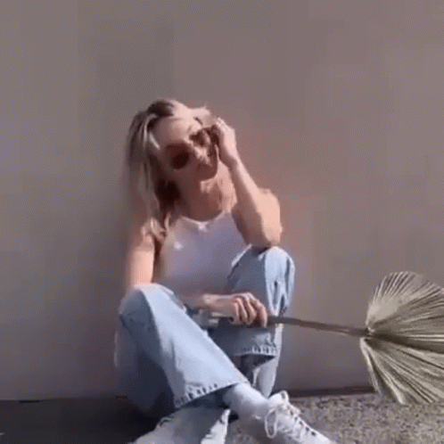 A Woman In A White Tank Top And Blue Jeans Is Sitting On The Ground With Her Legs Crossed . GIF