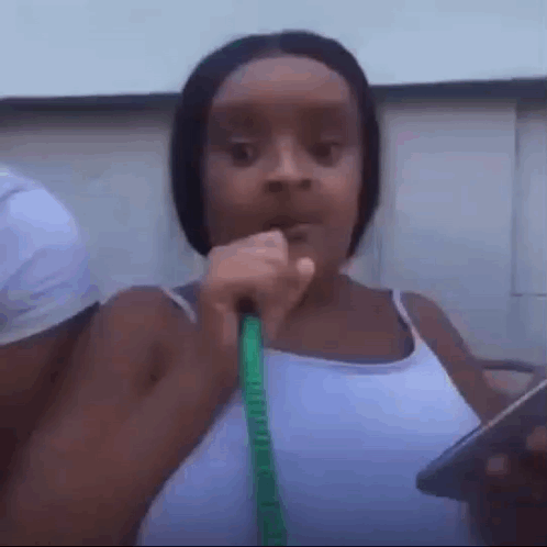 A Woman In A White Tank Top Is Holding A Green Hose In Her Mouth . GIF