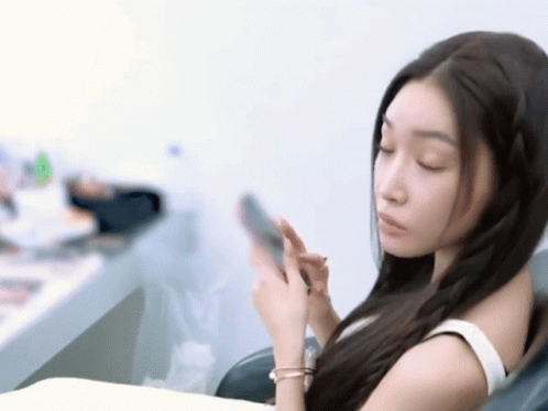 A Woman In A White Tank Top Is Looking At Her Phone GIF