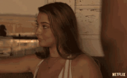A Woman In A White Tank Top Is Sitting On A Couch And Looking At Something . GIF
