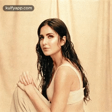 A Woman In A White Tank Top Is Sitting On The Floor With Her Legs Crossed And Looking At The Camera . GIF