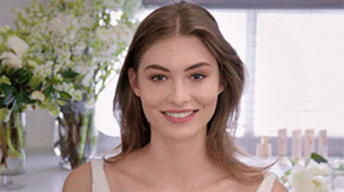 A Woman In A White Tank Top Is Smiling In Front Of Flowers . GIF