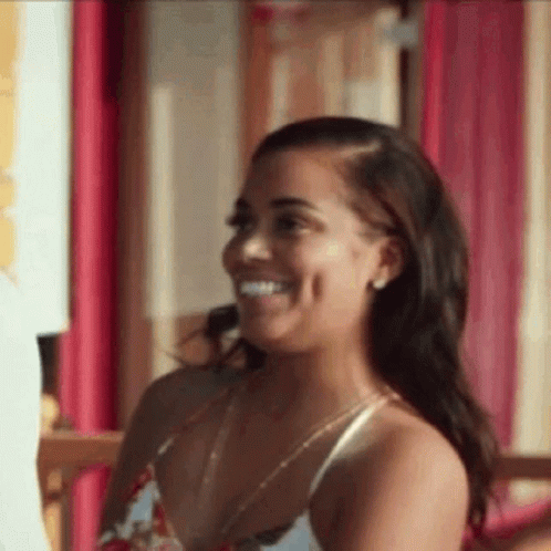A Woman In A White Tank Top Is Smiling While Sitting In Front Of A Pink Wall . GIF