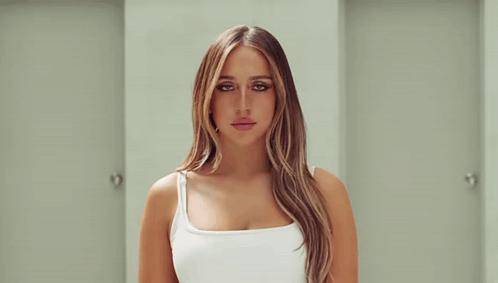 A Woman In A White Tank Top Is Standing In A Hallway Between Two Doors . GIF