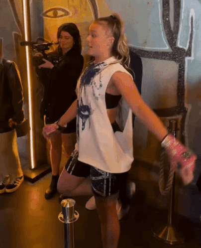 A Woman In A White Tank Top Is Standing In Front Of A Camera GIF