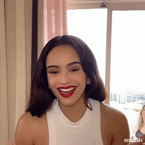 A Woman In A White Tank Top Smiles In Front Of A Window With Rosaliagifs Written Below Her GIF