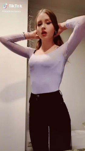 A Woman In A White Top And Black Pants Is Standing In Front Of A Wall With Her Hands Behind Her Ears . GIF