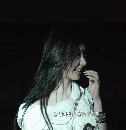 A Woman In A White Top Is Smiling In A Dark Room With Korean Writing On The Bottom GIF