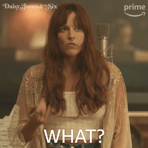 A Woman In A White Top Says " What " While Standing In Front Of A Microphone GIF