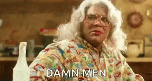 A Woman In A Wig And Glasses Is Sitting At A Table In A Kitchen And Says `` Damn Men '' . GIF