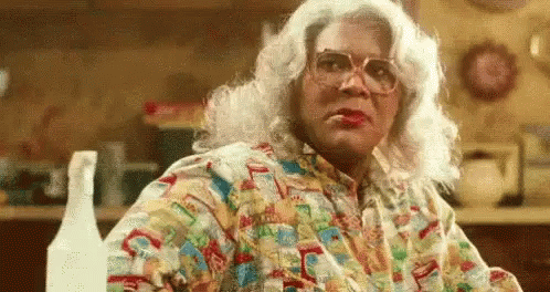 A Woman In A Wig And Glasses Is Sitting At A Table In A Kitchen With A Spray Bottle . GIF