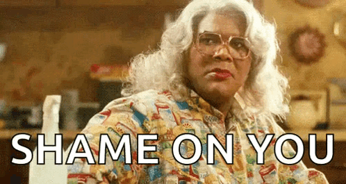 A Woman In A Wig And Glasses Is Sitting At A Table With The Words `` Shame On You '' Written On It . GIF