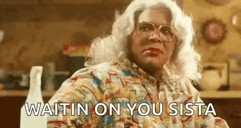 A Woman In A Wig And Glasses Is Sitting At A Table With The Words `` Waitin On You Sista '' . GIF