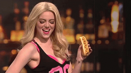 A Woman In A Wig Is Holding A Hot Dog That Says ' Vava ' On It GIF