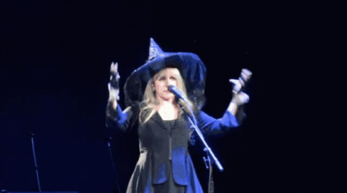 A Woman In A Witch Hat Sings Into A Microphone GIF