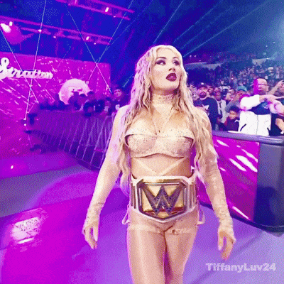 A Woman In A Wrestling Costume With A W Belt Is Walking On A Stage . GIF
