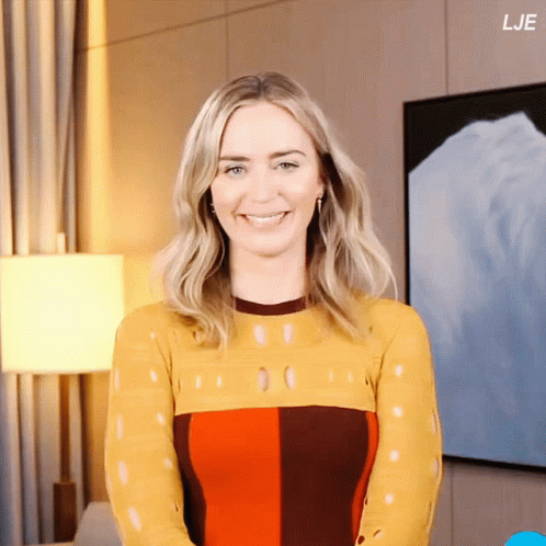 A Woman In A Yellow And Red Dress Smiles In Front Of A Wall With Lje On It GIF