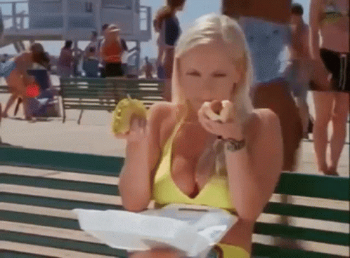 A Woman In A Yellow Bikini Is Sitting On A Bench Eating A Donut GIF