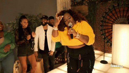 A Woman In A Yellow Crop Top Singing Into A Microphone In Front Of People GIF